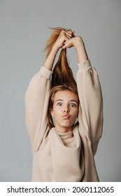 Funny grimacing teenage girl pulling her hair up, looking at the camera, making kissing lips. Against grey background. In a warm beige longsleeve.
