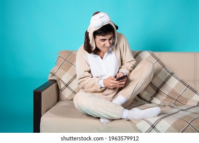 A Funny Guy Is Playing On The Phone While Sitting On The Couch. Blue Background. Young Man In Pajamas.