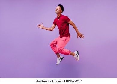 Funny guy in red t-shirt jumping and looking up. Studio portrait of emotional african male model posing on purple background.