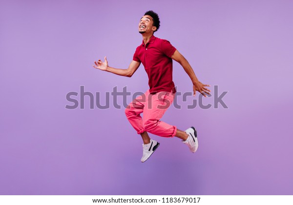 赤いtシャツを着たおかしな男がジャンプして上を見上げる 紫の背景にポーズを設定する感情的なアフリカの男性モデルのスタジオポートレート の写真素材 今すぐ編集