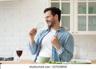 Funny Guy Singing Song Holding Beater Whisk Microphone Listening Music In Modern Kitchen, Happy Joyful Young Man Having Fun Cooking Dancing Preparing Healthy Dinner Meal Drink Wine Alone At Home