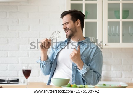 Funny guy singing song holding beater whisk microphone listening music in modern kitchen, happy joyful young man having fun cooking dancing preparing healthy dinner meal drink wine alone at home