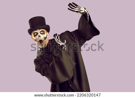 Funny guy in skeleton Halloween costume is scared of something. Man in black top hat, gloves and cloak, with skull makeup does protecting hand gesture and looks away with scared surprised expression