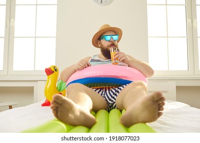 Funny Guy Spends Summer Vacation Inside His Apartment Due To Covid Pandemic Lockdown. Barefoot Chubby Young Man In Inflatable Lifebuoy And Beach Holiday Sunglasses Sipping Juice In Quarantine At Home