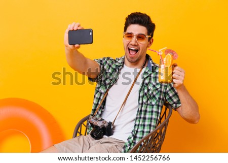 Funny guy in stylish shirt and white t-shirt makes selfie with glass of orange cocktail and winks on isolated background