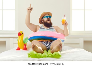 Funny guy in sunglasses, sun hat and swim ring sitting on beach mattress and sipping cocktail points finger up struck by cool idea on how to turn quarantine holiday at home into fun summer vacation