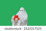 Funny halloween ghost in white sheet holding gift box, happy expression, isolated on chroma key green screen background. Happy halloween spooky concept.