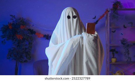 Funny halloween ghost in white sheet with black eyes in dark room talking on smartphone by video call, positive expression. Happy halloween spooky concept. - Powered by Shutterstock