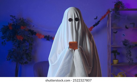 Funny halloween ghost in white sheet with black eyes in dark room texting on smartphone, sending message in app. Happy halloween spooky concept. - Powered by Shutterstock