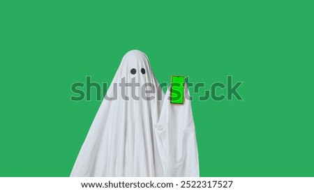 Funny halloween ghost in white sheet pointing shows thumb up at smartphone isolated on chroma key green screen background. Happy halloween spooky concept.