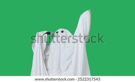Funny halloween ghost in white sheet singing son in karaoke microphone, isolated on chroma key green screen background. Happy halloween spooky concept.