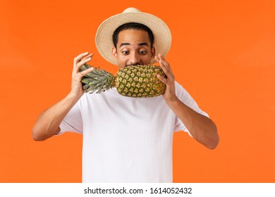 Funny handsome hipster guy, african american tourist enjoying vacation, trying bite pineapple, looking at fruit, standing orange background joyful, like travel hot countries during winter season