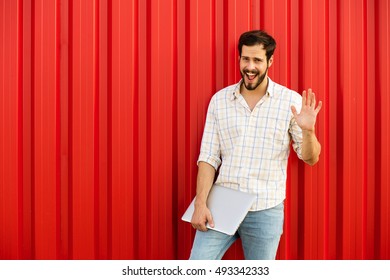 Funny Handsome Man Waveing Hand To Camera With Laptop On The Other Hand, Outside On A Red Background