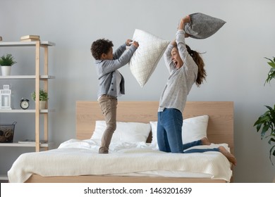 Funny Happy African Mixed Race Ethnicity Family Mum And Little Cute Son Having Fun Pillow Fight On Bed, Young Mother Laughing Playing Funny Game Enjoy Leisure Activity With Small Child Boy In Bedroom