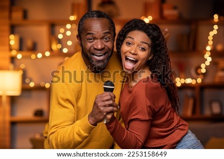 Funny happy black couple enjoying singing karaoke at home, holding microphone and singing songs together, closeup shot, living room decorated with lights interior. Leisure, entertainment concept