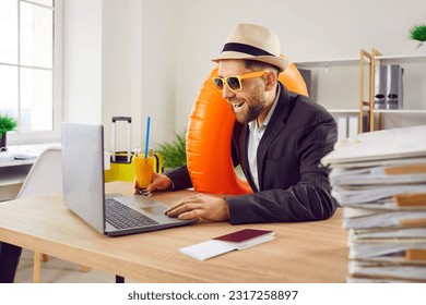 Funny happy business man sitting at the desk of his workplace in office with juice cocktail, boarding pass and passport holding rubber ring and booking tickets for summer vacation online via laptop.
