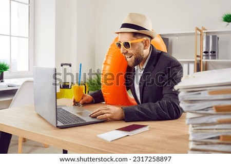 Funny happy business man sitting at the desk of his workplace in office with juice cocktail, boarding pass and passport holding rubber ring and booking tickets for summer vacation online via laptop.