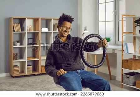 Funny, happy, confident African American man in glasses sitting on a chair in the living room at home, holding a black leather steering wheel cover and pretending to drive an invisible, imaginary car