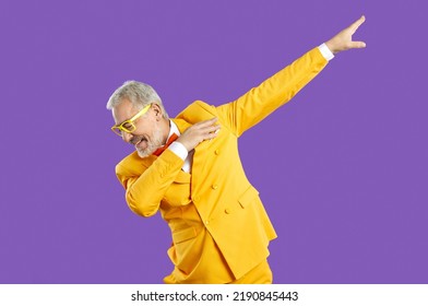 Funny happy confident handsome senior man in yellow suit and eyeglasses doing dab moves. Cheerful eccentric mature guy wearing trendy outfit dancing isolated on purple background. Fashion concept