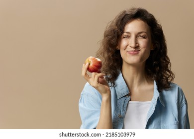 Funny happy cute curly beautiful female in jeans casual shirt chewing enjoy apple posing isolated on over beige pastel background. Healthy food. Natural eco-friendly products concept. Copy space