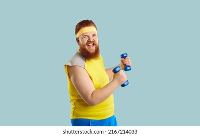 Funny happy excited bearded redhead plump fat man in yellow top and sweatband having fun while doing sports exercises with dumbbells. Studio shot, isolated on blue background. Training workout concept