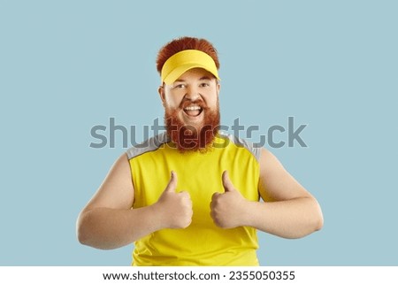 Funny happy fat man showing thumbs up motivating you to exercise isolated on light blue background. Young red-haired bearded man in sporty visor and T-shirt looks at camera with funny expression