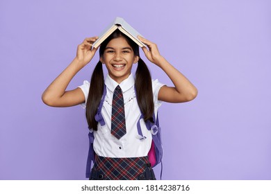Funny happy indian child primary school girl laughing holding open book over head. Positive cute smart latin kid schoolgirl with ponytails wearing uniform and backpack isolated on violet background.