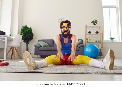 Funny happy inflexible man in glasses, retro sportswear and sweatband trying to do wide angle seated forward bend on floor at home. Fitness motivation, exercising, stretching, improving flexibility