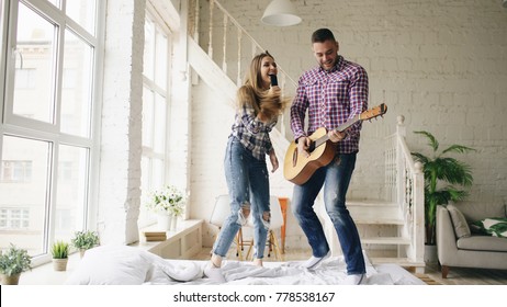 Funny Happy And Loving Couple Dance On Bed Singing With Tv Controller And Playing Guitar. Man And Woman Have Fun During Their Holiday At Home