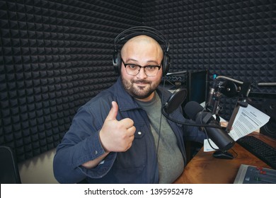 Funny happy radio presenter or host in radio station studio looks at camera and shows thumbs up gesture, portrait of cheerful working man.