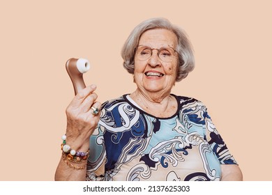 Funny happy smiling elderly grandmother holding satisfyer dildo with hand at studio over beige background. Senior old woman looking at vibrator with funny face expression.