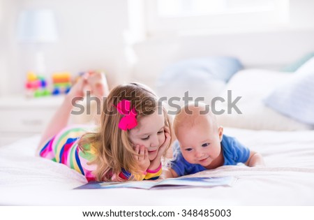 Funny happy toddler girl reading a book and playing with newborn baby boy in bed. Kids play at home. White nursery. Child in sunny bedroom. Children read and study. Interior for baby and young kid.