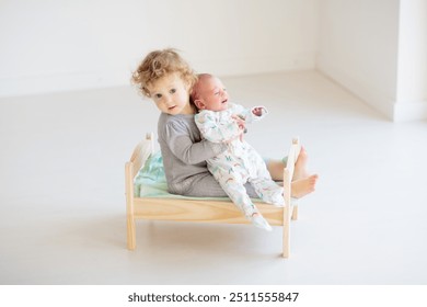 Funny happy toddler playing with newborn baby boy in toy bed. Kids play at home. White nursery for brother and sister. Child in sunny bedroom. Children meet new sibling. Interior for baby and kid. - Powered by Shutterstock