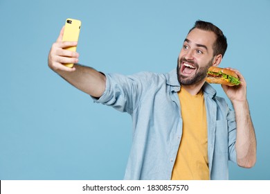 Funny Happy Young Bearded Man 20s In Casual Clothes Posing Holding In Hands American Classic Fast Food Burger Doing Selfie Shot On Mobile Phone Isolated On Pastel Blue Color Background Studio Portrait