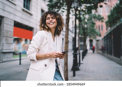 Funny hipster girl in headphones enjoying leisure time with positive music playlist for city walking during weekend, emotional female generation Z with mobile phone in hand smiling at streets