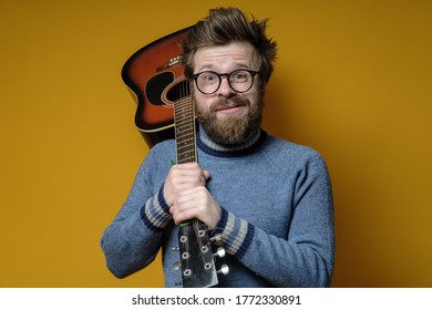 Funny Hipster In An Old Sweater Holds An Acoustic Guitar On Shoulder And Looks At The Camera With A Strange Grimace. Hobbies, Lifestyle. Studio, Yellow Background.