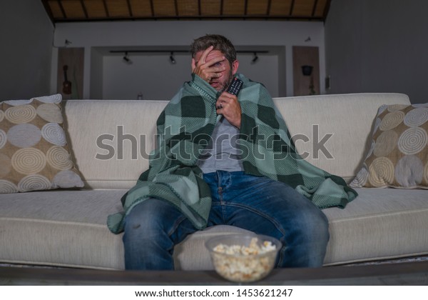 funny home\
lifestyle portrait of scared and frightened man alone at night in\
living room couch watching horror scary movie or scary show in\
television eating popcorn covering his\
face