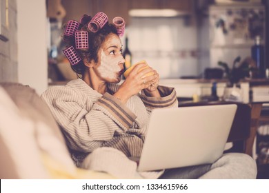 Funny housewife at home with pink curlers drinking a cup of tea while use a laptop on the sofa - technology and daily lifestyle at home - vintage filter colors and middle age lady enjoying the indoor 