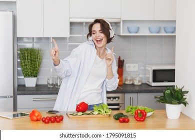 Funny housewife woman in casual clothes preparing vegetable salad cooking food in light kitchen at home. Dieting healthy lifestyle concept. Listen music with headphones, dancing fooling around 