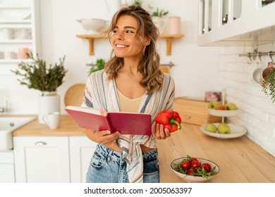 Funny Housewife Woman Looking At Recipe In Cookery Book Preparing Vegetable Salad Cooking Food In Light Kitchen At Home. Dieting Healthy Lifestyle Concept.