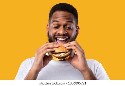 Funny Hungry Black Guy Biting Burger Eating Junk Food Standing Over Yellow Studio Background. African Man Enjoying Hamburger. Unhealthy Nutrition And Binge Eating Habit