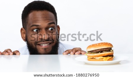 Funny Hungry Black Man Looking At Burger On Table Posing Over White Studio Background. Nutrition And Dieting, Cheat Meal And Unhealthy Junk Food, Overeating Habit Concept. Panorama
