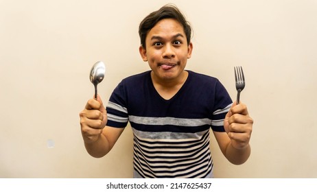 Funny And Hungry Face Expression Of Young Asian Malay Man With Strips T-shirt Holding A Spoon And Fork Ready To Eat. Licking Lips, Want To Eat, Diet And Healthy Food Concept. Selective Focus.