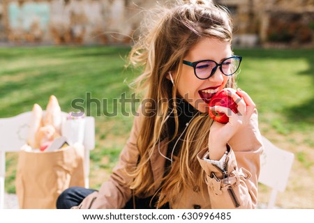 Funny hungry girl with appetite bites off red apple sitting in the park after a shopping trip. Attractive young woman eating turning away on the blur nature background. Lunch in the park, buying food