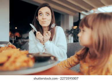 
Funny Hungry Mom Holding Knife and Fork. Candid moment of a family having lunch in a restaurant
