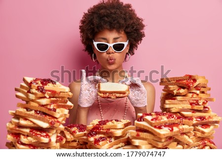 Funny hungry woman eats tasty bread sandwich, keeps lips rounded, dressed elegantly, wears trendy sunglasses, enjoys tasty lunch, has good appetite and gluttony, comes closely to pile of toasts