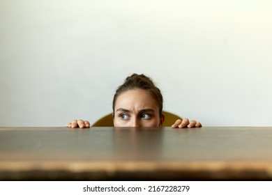 Funny indoor picture of frowning student girl with hair knot and guarded look hiding behind table, concentrated eyes looking aside, listening attentively to strange sounds and noise