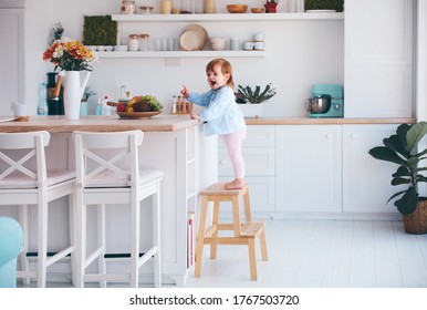 Funny Infant Baby Girl Standing On A Step Stool At The Cozy Kitchen At Home