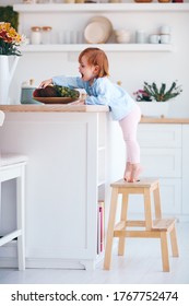 Funny Infant Baby Girl Standing On A Step Stool At The Cozy Kitchen At Home