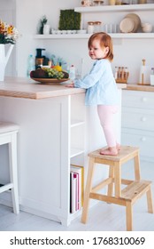 Funny Infant Baby Girl Standing On A Step Stool At The Cozy Kitchen At Home